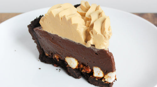 A single slice of Rocky Road Pie on a white plate, showing distinct layers: a chocolate cookie crust, a rocky road base with marshmallows and smoked almonds, a dark chocolate filling, and a fluffy Salted Caramel Whipped Cream topping.